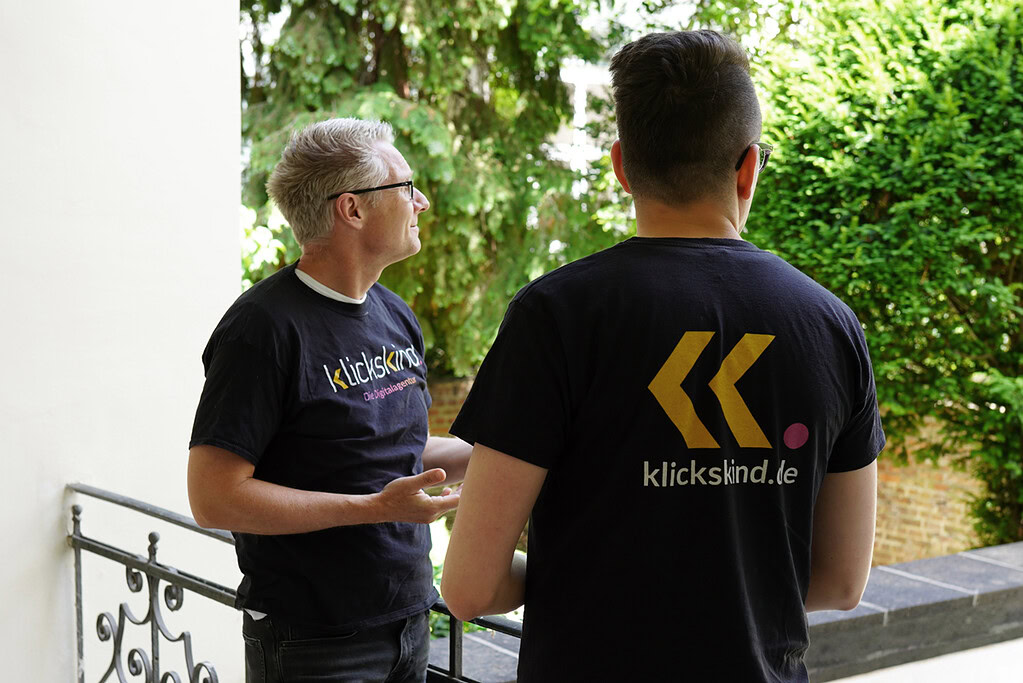 Zwei Mitglieder des klickskind-Teams unterhalten sich draußen und tragen schwarze klickskind-Shirts.